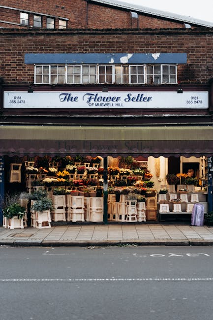A floral shop named 'The Flower Seller of Muswell Hill' with a dark awning and a large window display showcasing a variety of fresh flower bouquets and floral arrangements. The display includes vibran