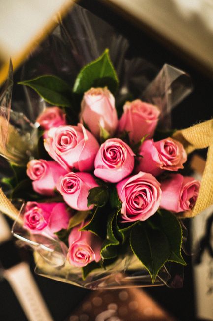 A bouquet of fresh pink roses arranged with dark green leaves, wrapped in clear plastic and tied with a yellow ribbon. The roses vary in size, with some fully opened, revealing delicate petals, while 