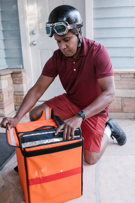 A male delivery person crouching on a porch with a neutral expression, preparing to deliver flowers. He is wearing an orange insulated delivery bag, a black helmet with goggles, a short-sleeved burgun