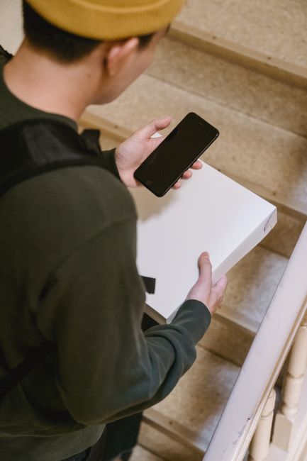 A person dressed in dark clothing and wearing a beige beret is seen holding a white floral gift box on a staircase. The individual is holding a black smartphone and appears to be either about to open 