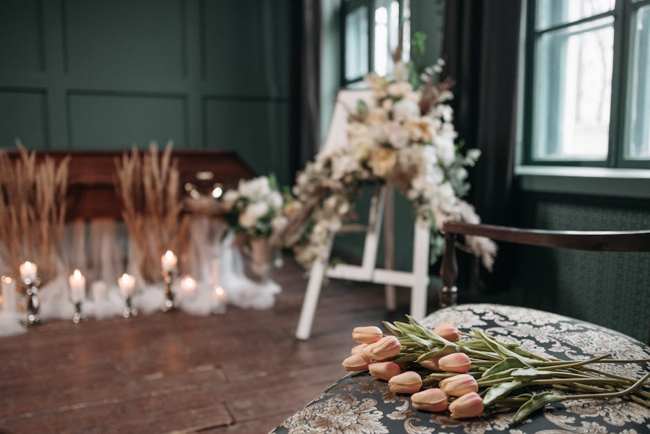 A floral arrangement featuring soft pink tulips with green stems and leaves resting on an ornate upholstered chair in a softly lit room. In the background, there is a large, elegant display of white f