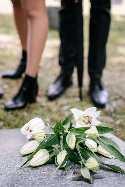 A bouquet of white lilies with large, open petals and prominent stamens, surrounded by unopened lily buds and glossy green leaves, resting on a stone surface outdoors. In the background, two people ar